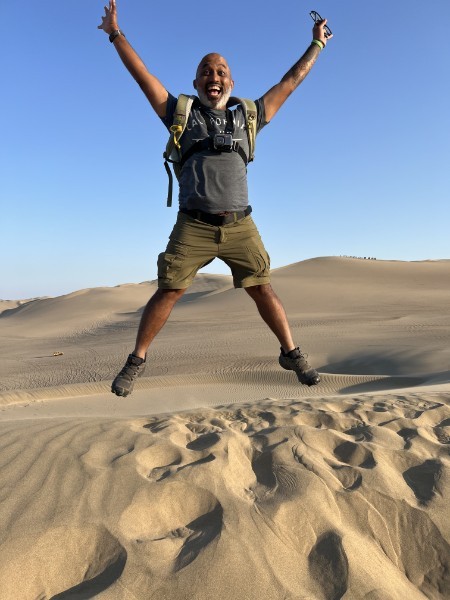 Sharif Rehman mid-jump with arms raised in the desert dunes, wearing a backpack and a chest-mounted camera